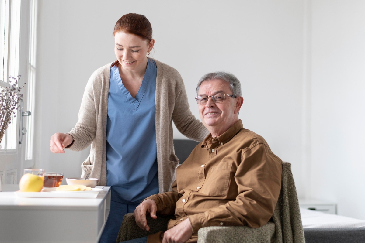 Apollo Beach (2) A cheerful female caregiver in scrubs and a cardigan provides assistance to a smiling elderly man seated at a table with tea and fruit, in a bright, cozy room—illustrating compassionate and professional home care in Apollo Beach.