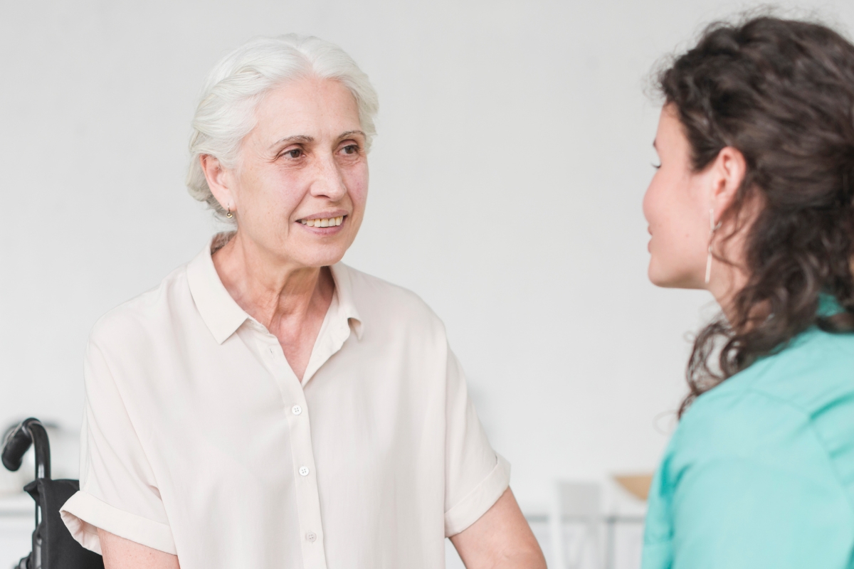 Bedford An elderly woman with white hair, seated next to a wheelchair, smiles warmly while engaging in conversation with a younger female caregiver. The scene reflects a calm, supportive atmosphere, symbolizing compassionate home care in Bradford focused on connection, dignity, and personalized support for seniors.