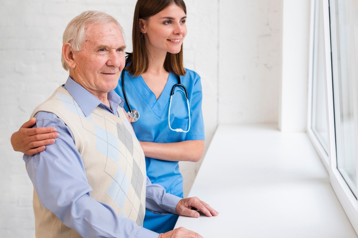 A young female caregiver in blue scrubs with a stethoscope gently supports an elderly man wearing a light sweater vest as they both look out a bright window. This image represents compassionate home care in Keystone, Florida, emphasizing trust, support, and companionship for seniors.