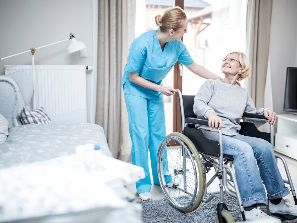 Home Care in North Dade A caregiver in blue scrubs assists a smiling senior woman in a wheelchair inside a bright, comfortable home setting, symbolizing compassionate home care in North Dade.