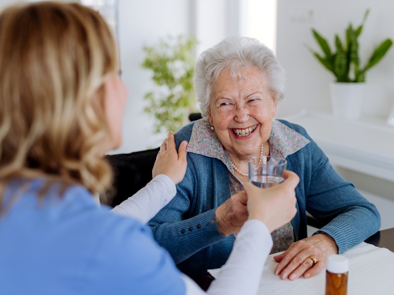 Warm and Joyful Medication Supervision in Temple Terrace A caring professional sharing a warm laugh and handing a glass of water to an older woman, providing gentle Medication Supervision Temple Terrace.